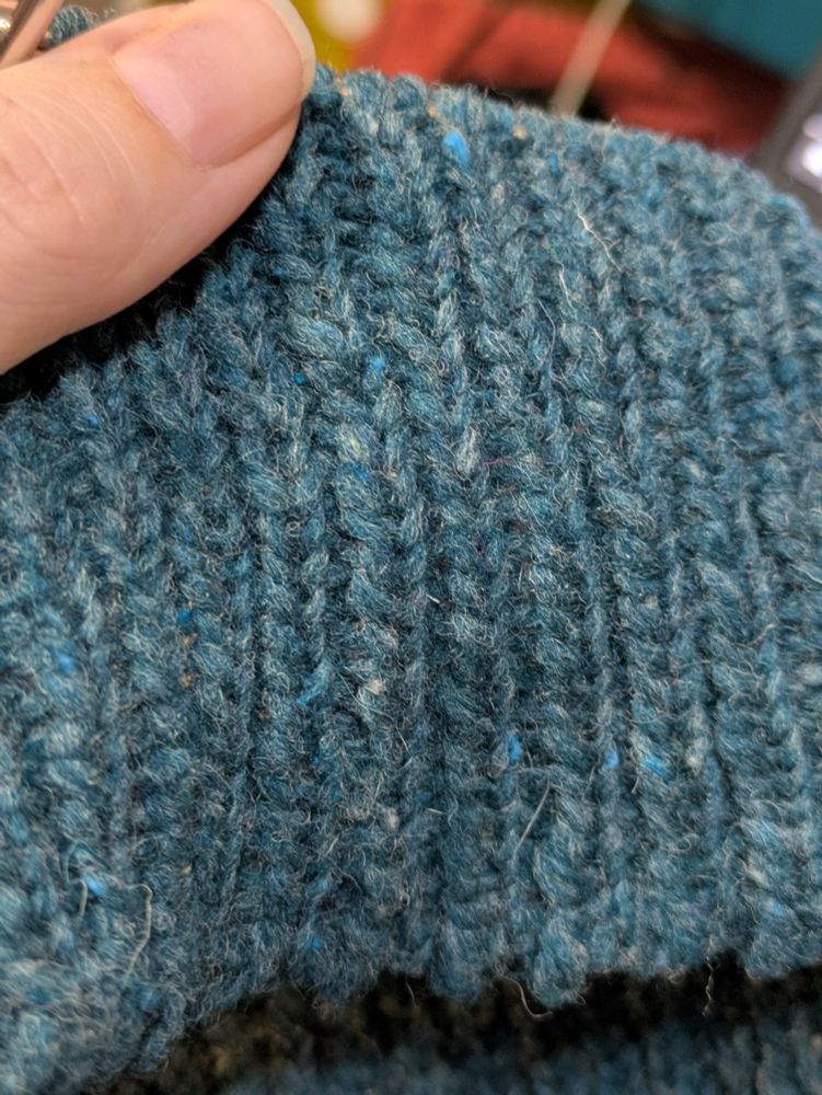 A close-up of one-by-one ribbing, worked in a slightly heathered, rustic yarn in a shade that might be teal or blue depending on the light and the camera. My left thumb is visible in the top corner of the picture, mainly as a marker of scale and a cheat for making my camera's colour balance more accurate (as a contrast to the yarn). 
The yarn looks pretty normal, all knit up. Maybe a little unevenly thick and thin, but nothing unusual. Before being knit, though, it's made of two parallel slender strands of unspun roving. 