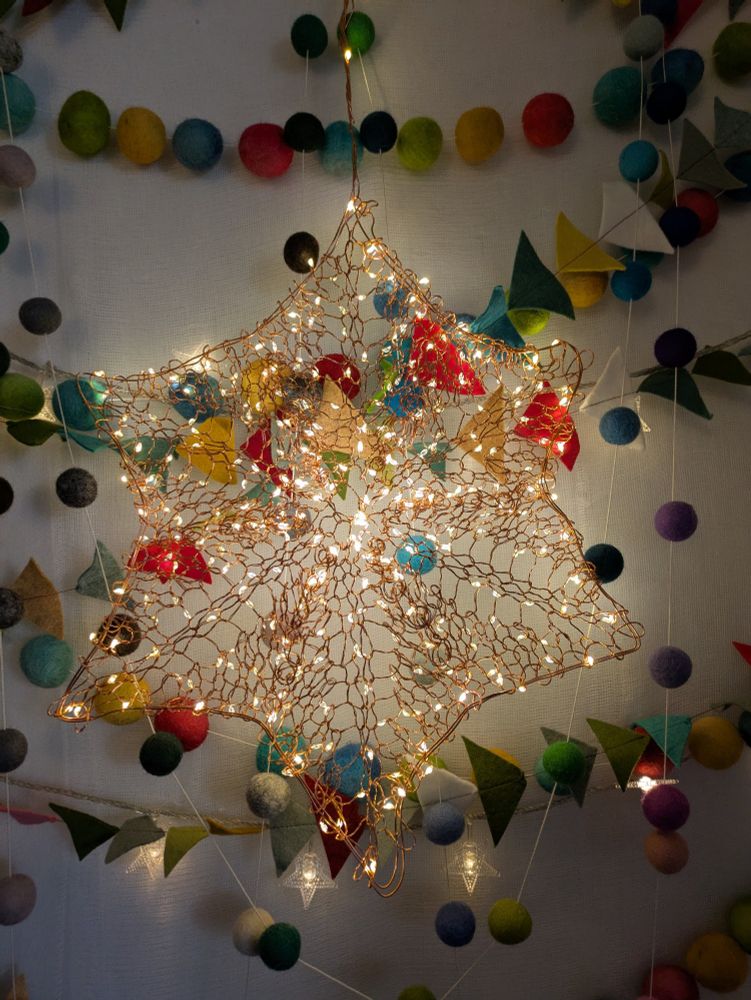 A 6-pointed star, knitted from the centre out with a long copper-wire strand of fairy lights. It's about 12" or 20cm tip to tip. It's hanging against a softly glowing backdrop: a layer of cheesecloth and more lights, and then layers of garlands. There's a garland made of triangles of multi-coloured felt, sewn together like a flying-geese quilt pattern; and there are at least two garlands made of rainbow coloured wool felt balls strung on white cotton yarn. The whole thing speaks of softness and warmth and light, and it's my sensory wall in my living room. 