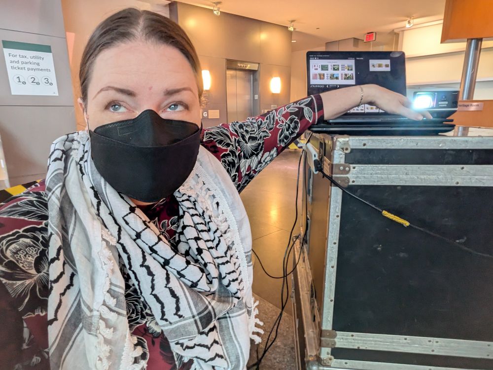 A white woman with brown hair and a black face mask, wearing a keffiyeh, has her arm raised awkwardly to press the arrow keys on a laptop that's on a tall cart beside her, a long with a projector. She's looking off camera to monitor what's on the projection screen. 