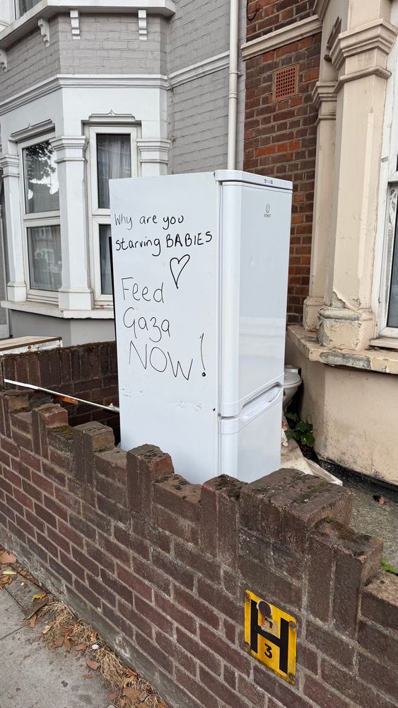 A white fridge freezer in someone’s garden. Written on the fridge in black marker are the words:
“Why are you starving babies Feed Gaza Now!”