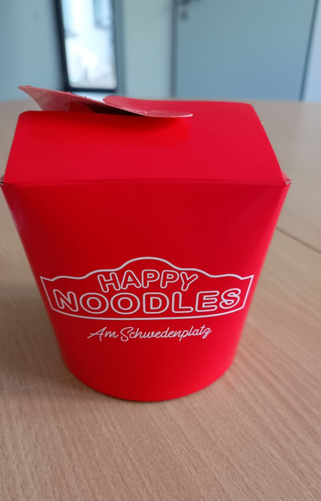 Caixa de comida chinesa de papel, cúbica, vermelha, em branco está escrito "Happy Noodles am Schwedenplatz"