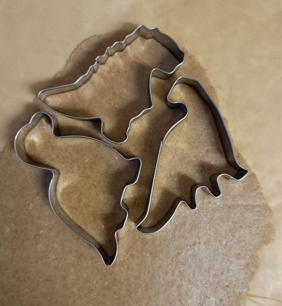 Massa de bolacha de natal com três cortadores metálicos em cima no formato de dinossauros. 