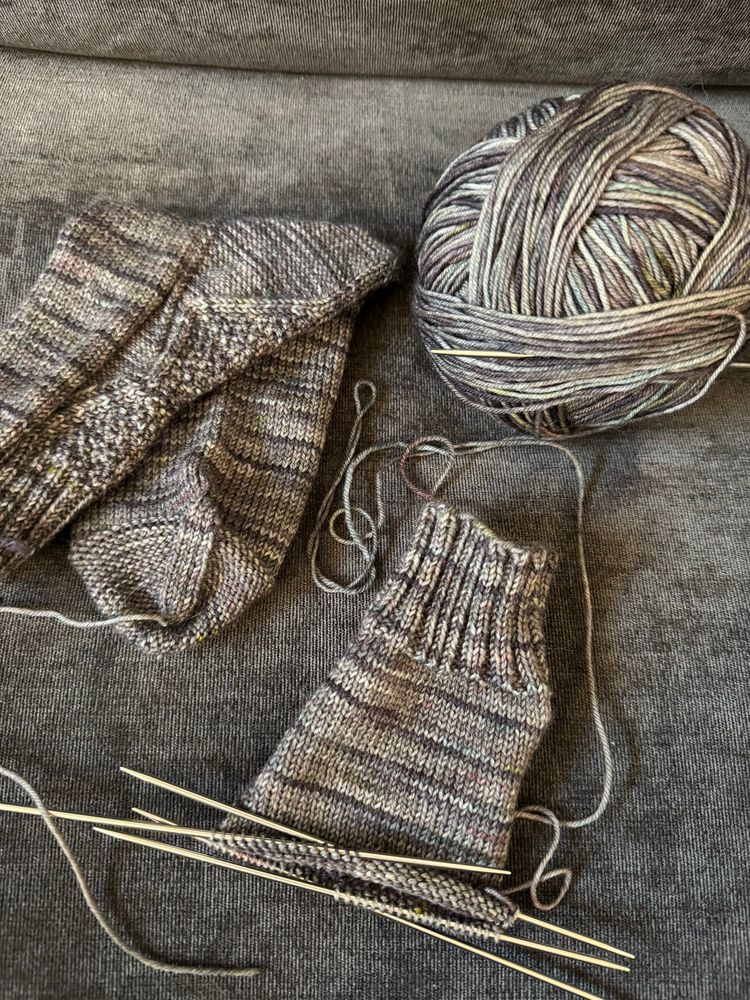 A finished sock, a partially knit sock and a ball of variegated grey yarn lay on a grey sofa cushion. 