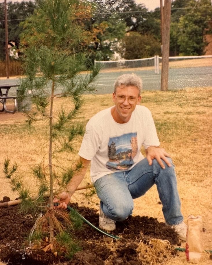 A white haired man crouching next to a tree he recently planted
