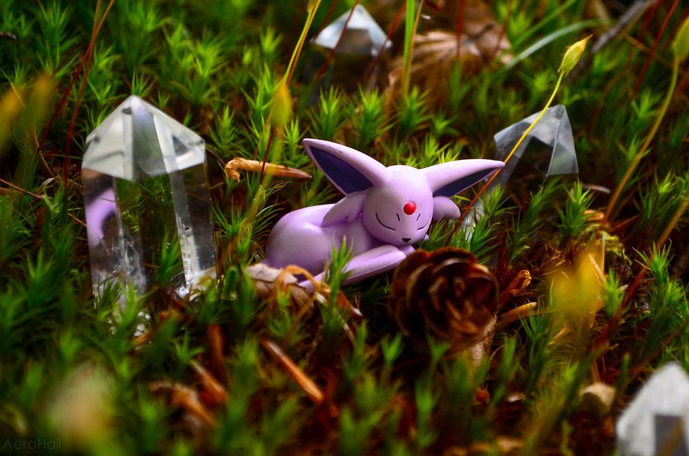 A photo of a tomy figure/toy of the pokemon Espeon sleeping in moss, there are crystal points distributed around the moss and a pinecone.