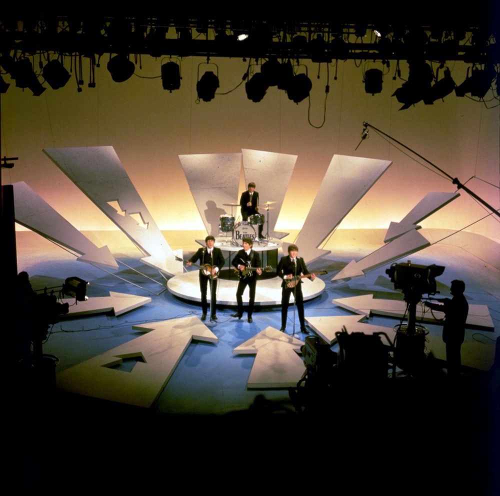 Vintage photo of the Beatles performing on the Ed Sullivan show in 1964. They play on a blue stage surrounded by white arrows, with a yellow cyclorama behind. Tv cameras flank them to the left and right. They wear black suits. 