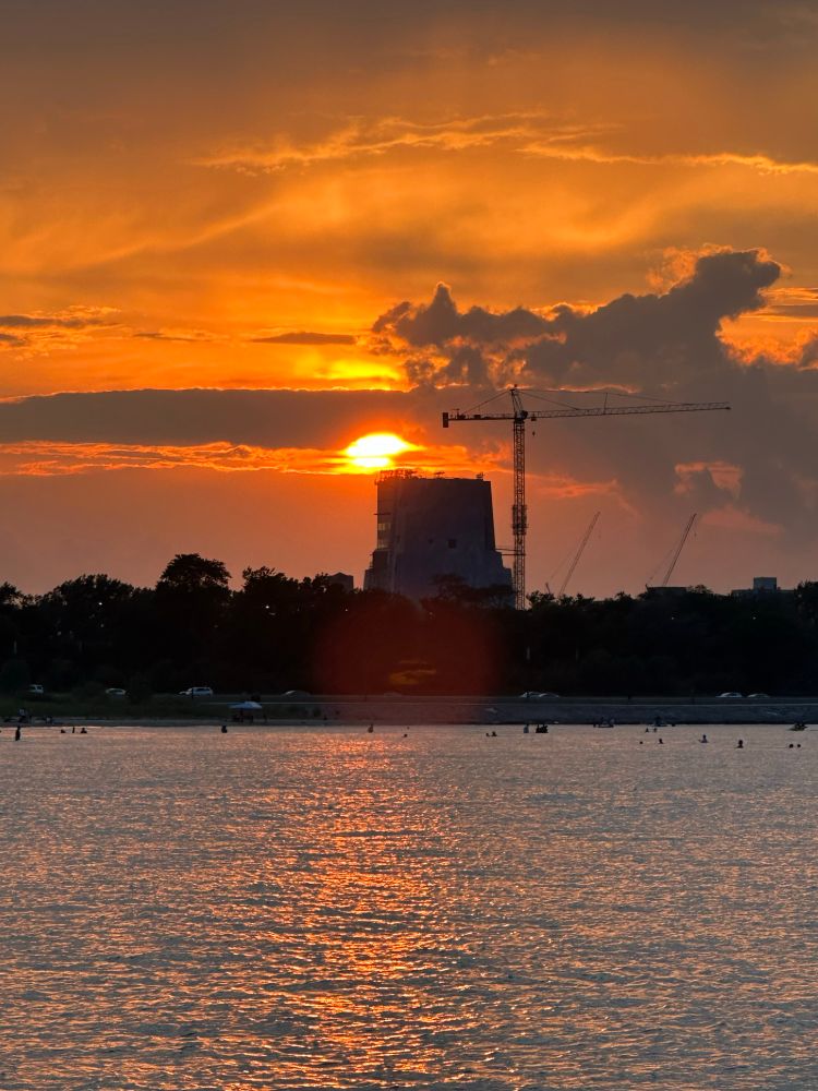 Tall building under construction with crane to one side and setting sun behind it. Sunset light reflects in the water of Lake Michigan