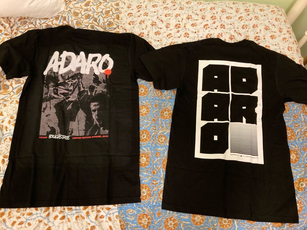 This image features two black T-shirts laid flat on a bed with a patterned bedspread. The T-shirt on the left shows the back design. It prominently features the name “Adaro” in bold white letters with a red dripping detail. Below this, there is a grayscale graphic collage of people and textures. At the bottom, it reads “Adaro,” “Roughstate,” and “Limited Edition Apparel 2K24” in red and white text. The T-shirt on the right shows the back design. It features six black blocks with bold white lettering that collectively spell “Adaro.” The bottom right block includes a striped design for added contrast. Beneath the blocks, smaller text reads, “Adaro - Limited Edition Apparel 2K24.” Both designs highlight a bold and modern aesthetic.
