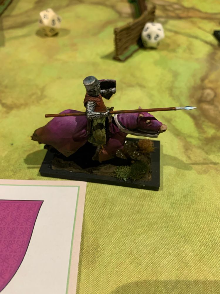 A painted knight figurine viewed from side on. He carries a lance and rides a brown horse clad in purple. 