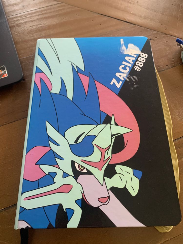 An A5 notebook. On the cover is Zacian, a wolf-like Pokémon with a sword in its jaws. 
