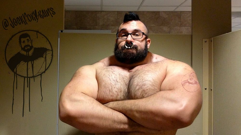 A digitally altered photo of a big muscular white man, with glasses, a mohawk and a thick black beard. He's flexing his muscular arms inside a stall.
His arms, chest and neck have been made bigger digitally.