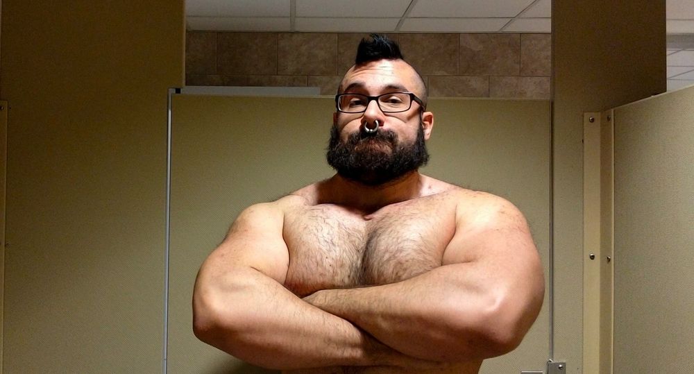 A big muscular white man, with glasses, a mohawk and a thick black beard. He's flexing his muscular arms inside a stall.