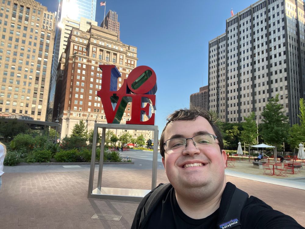 A selfie of the poster (a mid-20s white male with dark brown hair and glasses) in a black shirt and backpack standing in front of the Philadelphia “LOVE” sign (red block letters saying LOVE with the LO on top and the O at a 45 degree angle up and to the right, sitting on top of an open silver metal frame). A mostly empty plaza, several buildings, and some short bushes and trees are visible in the background. 