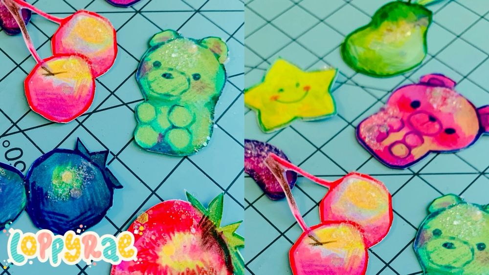 Small paper cutouts of blueberries, cherries, a strawberry, a pear, a star, and gummy bears drawn with bright colors and dipped in clear glitter.