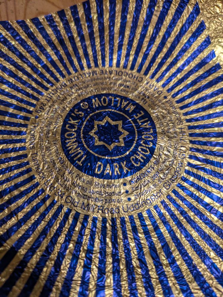 Tunnocks tea cake wrapper, dark chocolate variant. A dark blue eight pointed star, surrounded by a gold eight pointed star, and series of gold and blue concentric circles, finally surrounded by gold and blue lines which would converge at the centre of the original blue star. 