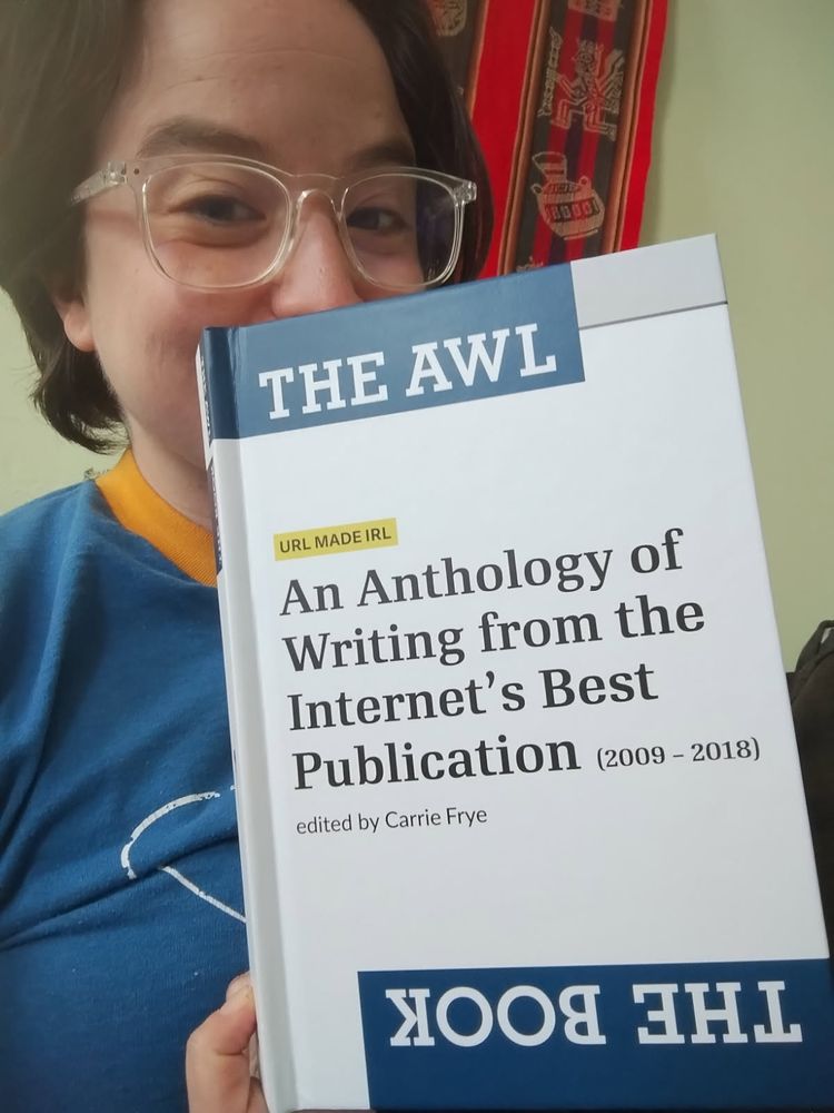 a brown haired extremely tired non profit salt miner with lucite reading glasses has their face behind a white book. The Cover is white with a blue square title reading: "THE AWL". Beneath it is a black title with, "An Anthology of Writing from the Internet's Best Publication (2009 - 2018), edited by Carrie Frye." They are very happy despite being very despondent.