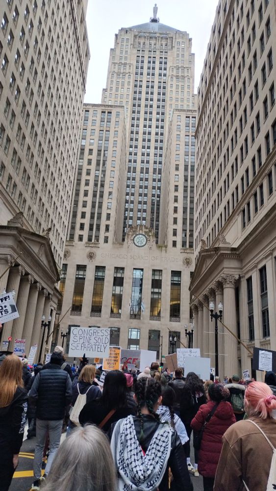 A crowd of protesters on a gray day with many signs in front of the beige limestone Chicago Board of Trade Building. It feels very prescient.