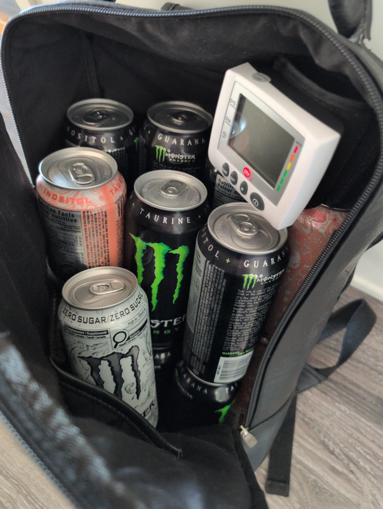~16 monster energy drinks of various flavours in a backpack with a blood pressure monitor.