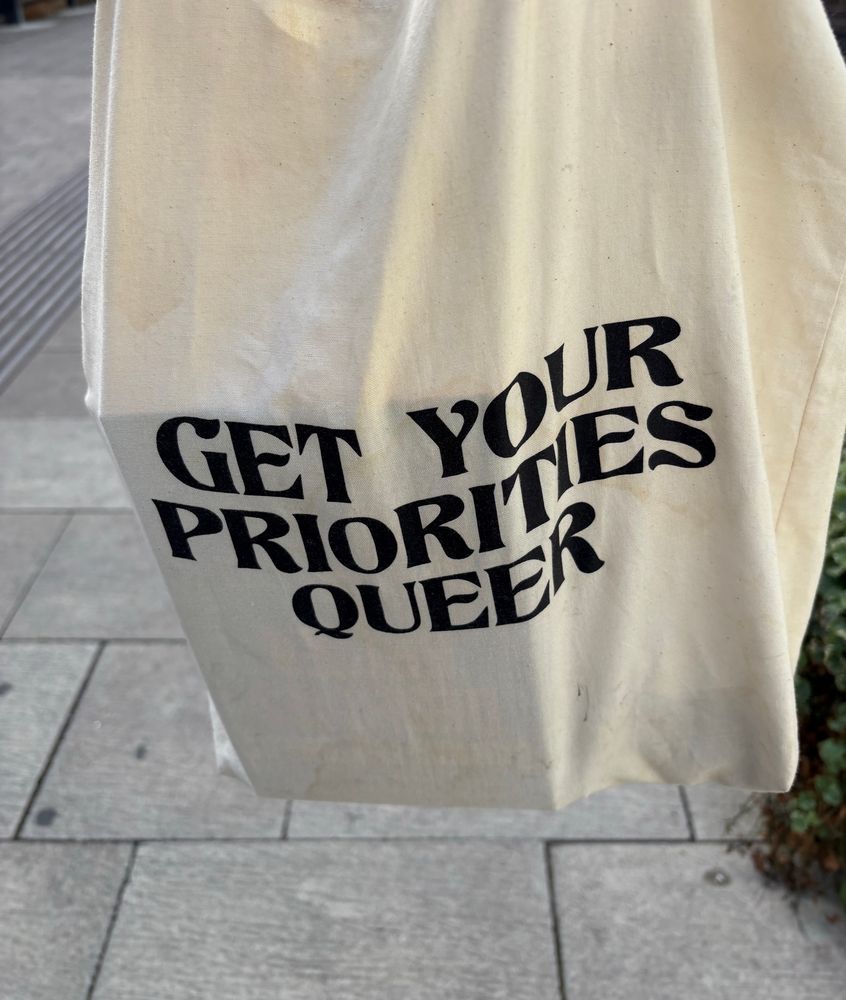 White tote bag with the text “get your priorities queer”.