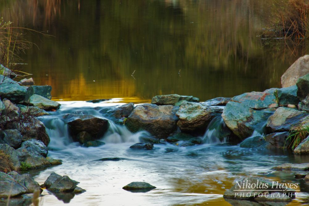 A series of rocks in a stream, forming a small waterfall. Reflections of green and yellow trees are seen in the water's surface.