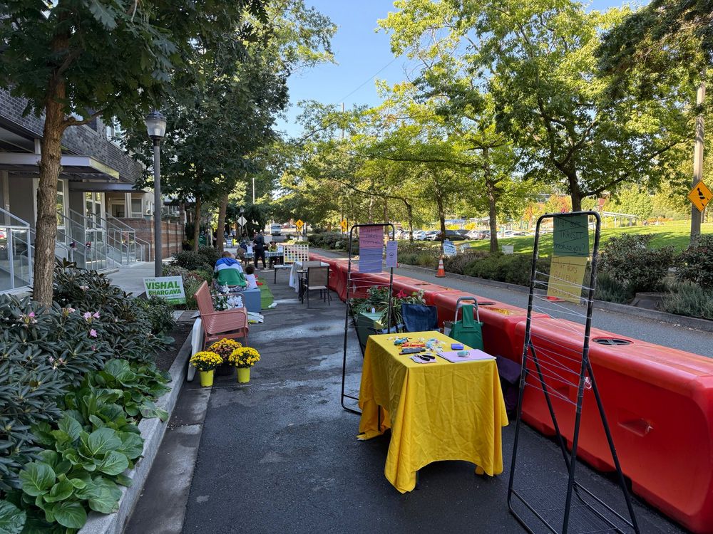 Picture of an outdoor area with tables, chairs, greenery, and people hanging out near a curb