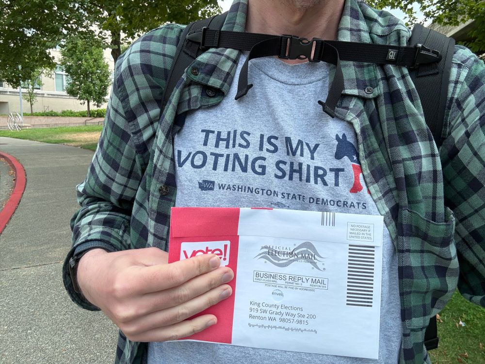 Picture of a King County elections ballot envelope held in front of a grey shirt that reads “this is my voting shirt, King County Democrats.”