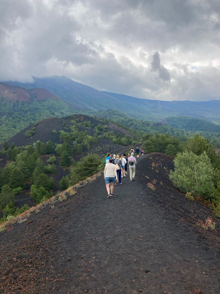 A group of people walk on hillocks of black ash. Smoking mountain in the background. 