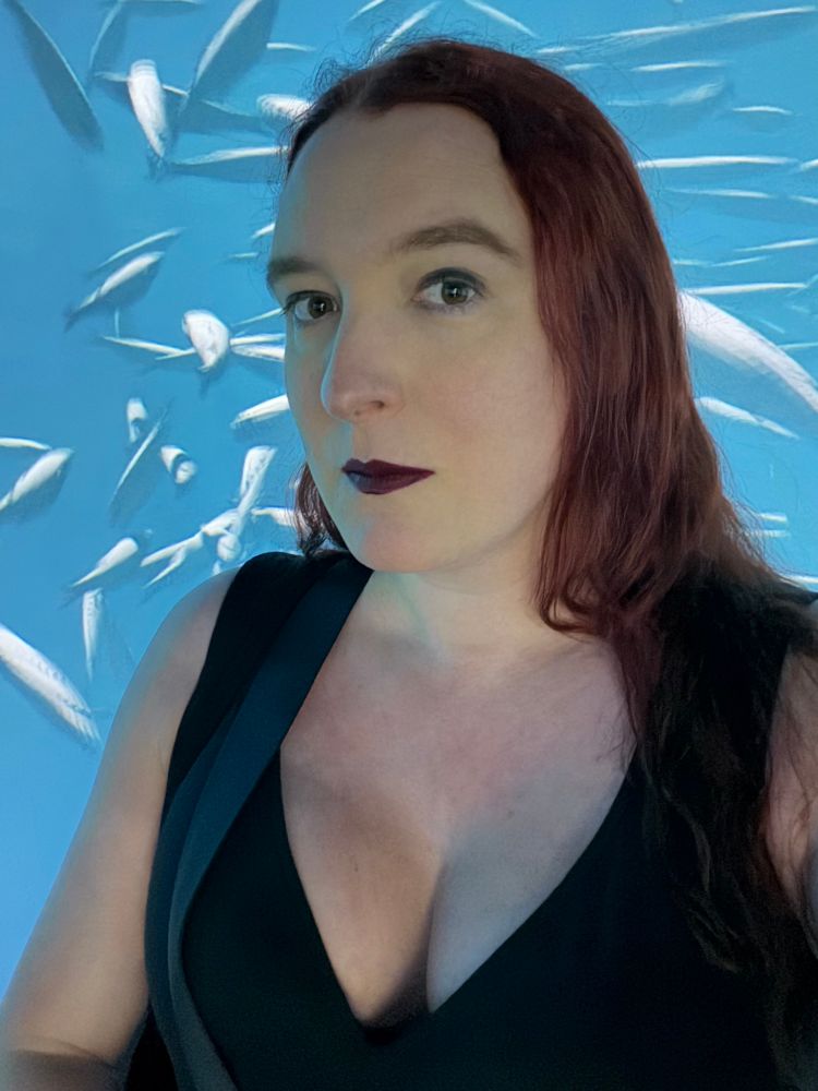 A curvy pale woman with fierce death femme makeup and a black dress. Behind her is a school of fish swimming, via digital projector.