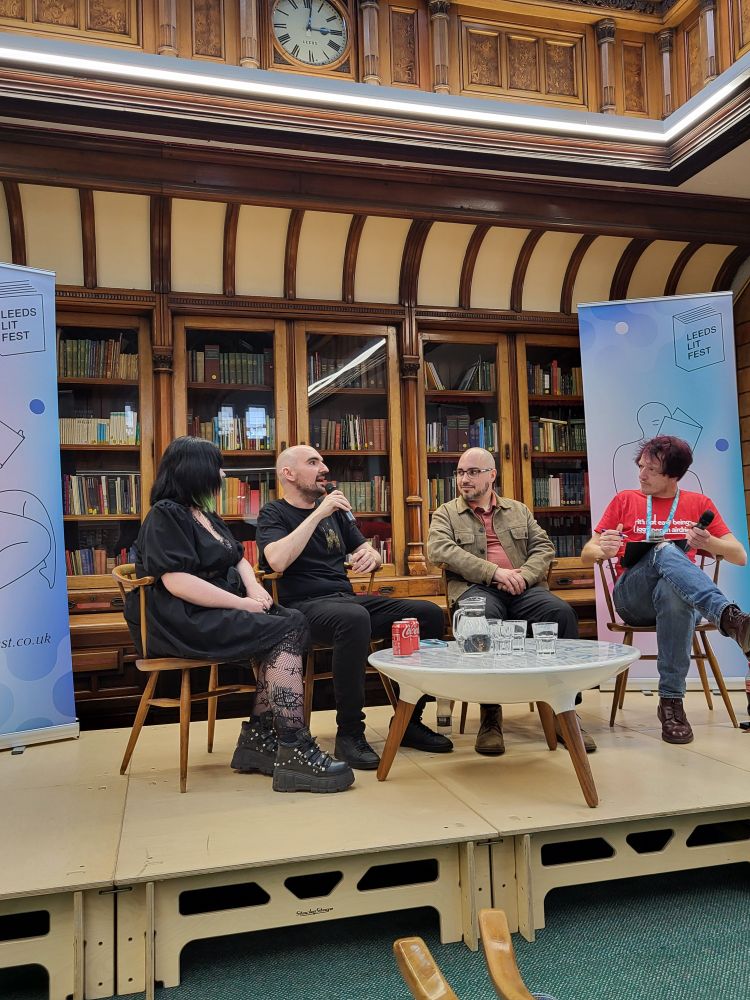 Amy, Ted, James from Northodox Press sat alongside Stu Hennigan in a panel event for Leeds Lit Fest. They are sat on raised platform with 2 festival banners on either side. 