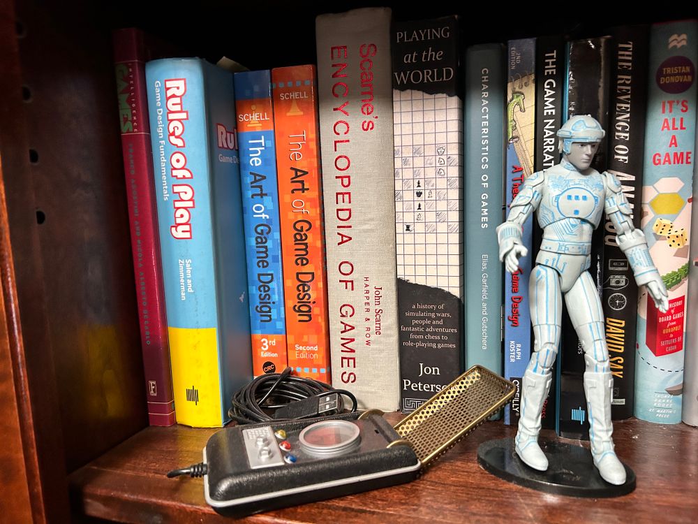 Left side of my game design library bookshelf with books like Rules of Play, Scarne’s Encyclopedia of Games, Playing at the World, and the Revenge of Analog. 