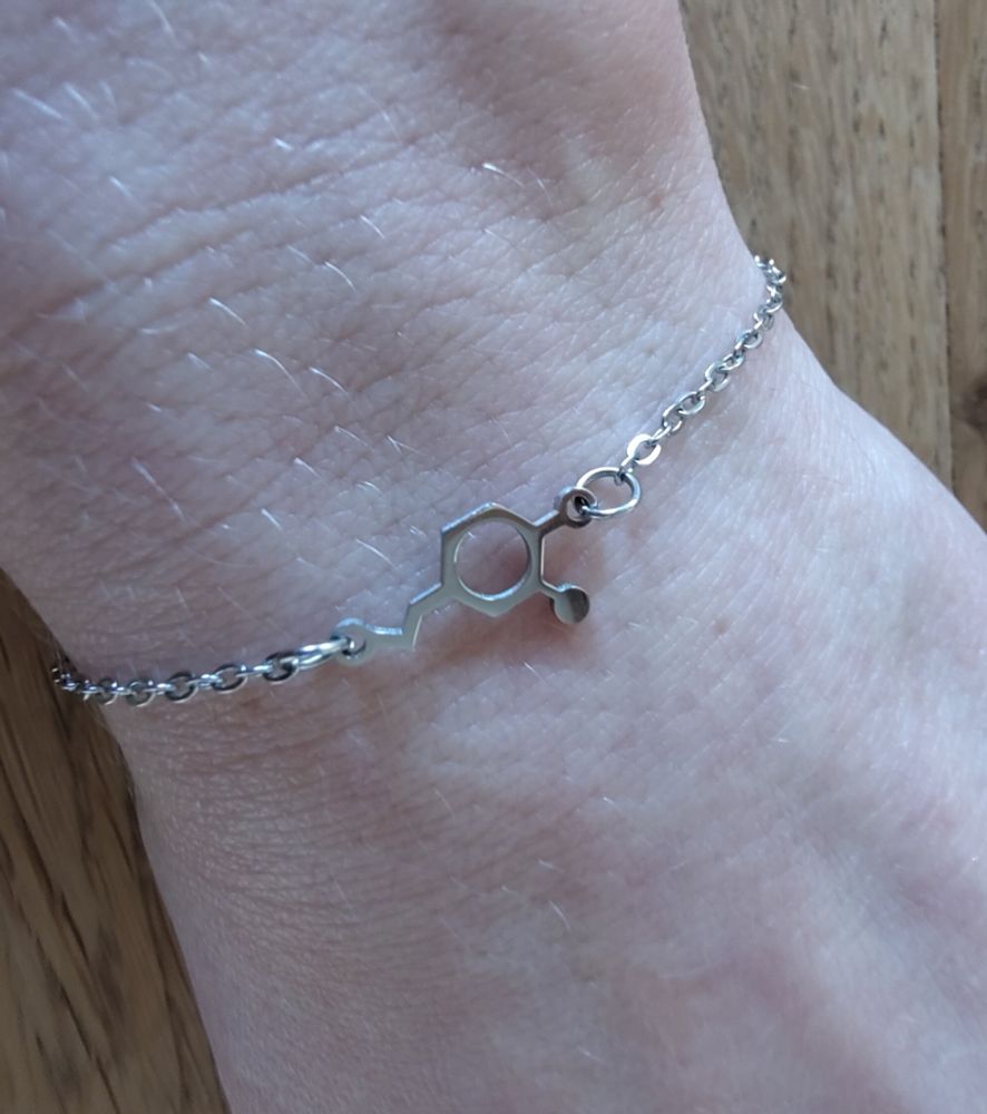 Ein Handgelenk weißer Hautfarbe mit einer silberfarbenen Kette mit der Molekülstruktur von Dopamin in der Mitte. Die Hand liegt auf einem Holztisch.