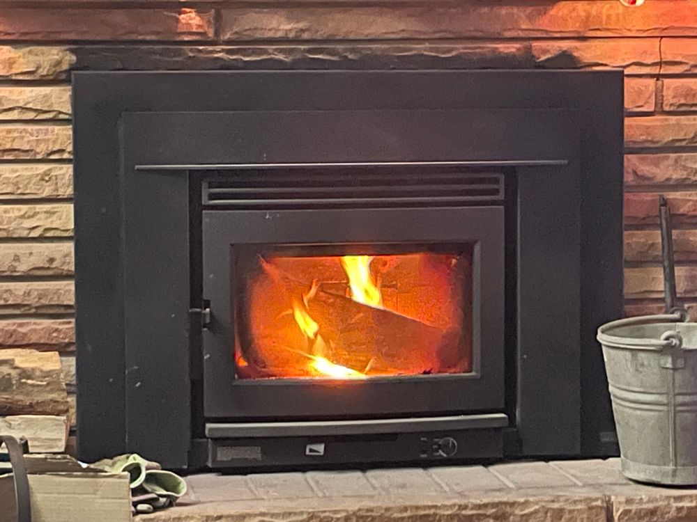 A fire is burning inside a black fireplace insert. The stones of rhe fireplace are shades of tan with hints of red. There is an ash bucket on the right side of the hearth, and gloves are visible laying to the left. 
