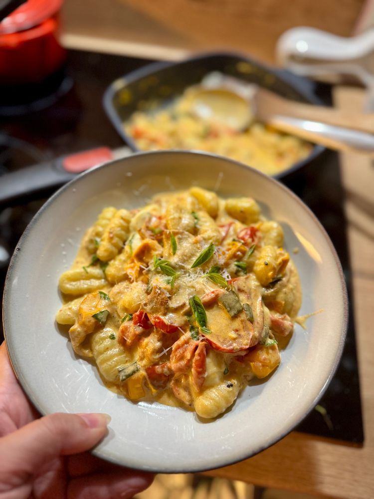 Das Bild zeigt eine Portion Gnocchi in einer cremigen Sauce mit gebratenen bunten Tomaten.  Die Gnocchi sind auf einem hellen Teller angerichtet und mit Basilikumblättern garniert.  Im Hintergrund ist unscharf ein weiteres Gericht in einer Pfanne zu sehen, das vermutlich die gleiche Zubereitung enthält.  Eine Hand hält den Teller.
