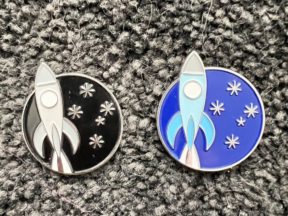 A pair of enamel pins/badges with the rocket and stars logo of Continuum 15. The one on the left is black, while the one on the right is blue.