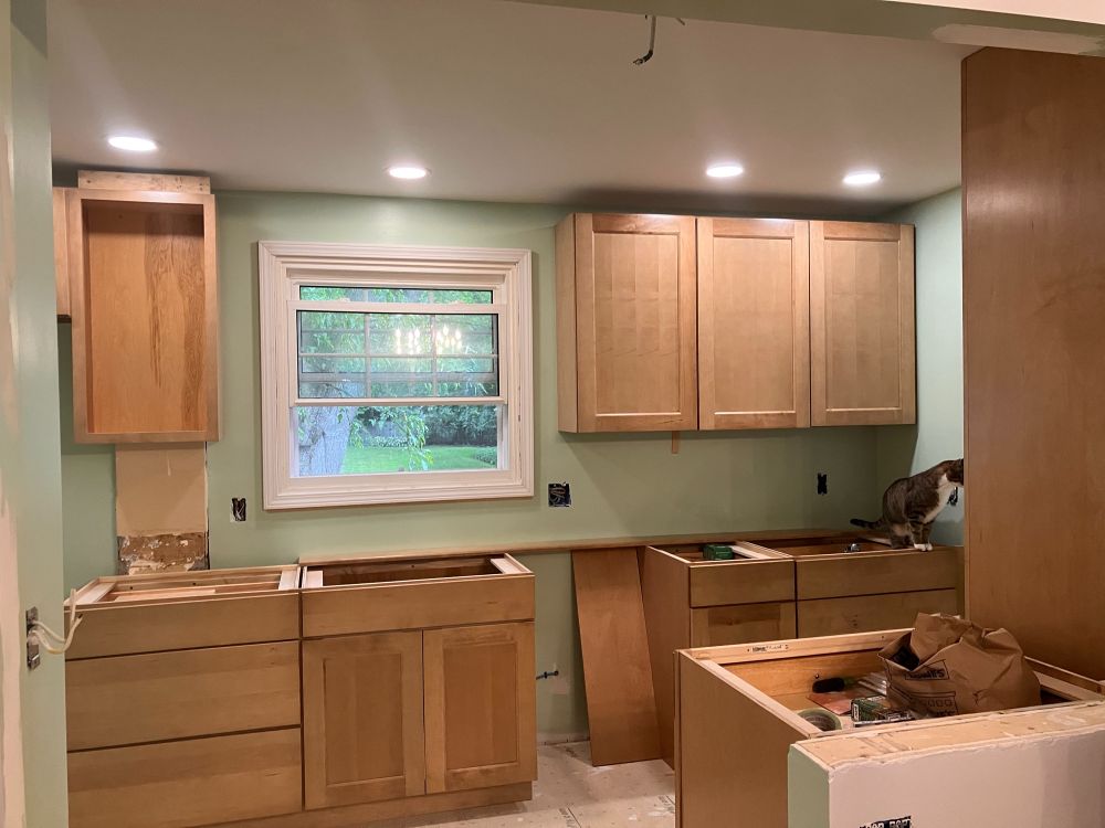 A photo of a kitchen in the process of being remodeled; light wood cabinets are set up without countertops or appliances, the walls are painted green, and most importantly: there are three dimmable lights in the ceiling now!