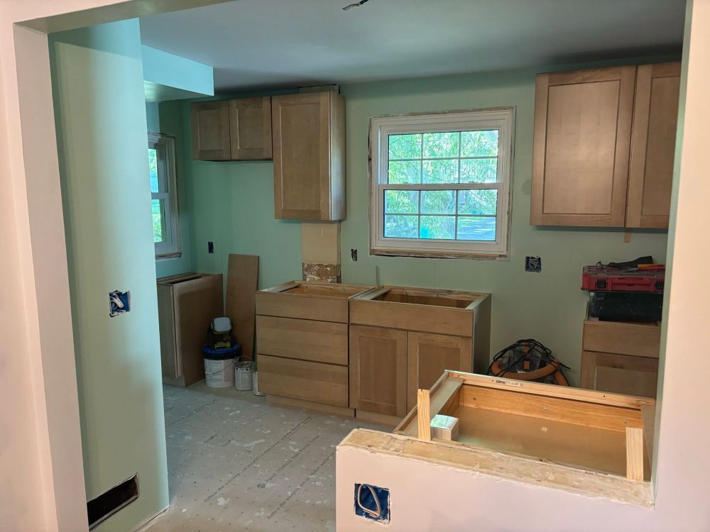 A photo of a kitchen in the process of being remodeled; the walls are painted green, the cabinets are a light wood and don’t yet have handles, and there are no countertops, flooring, or appliances. 