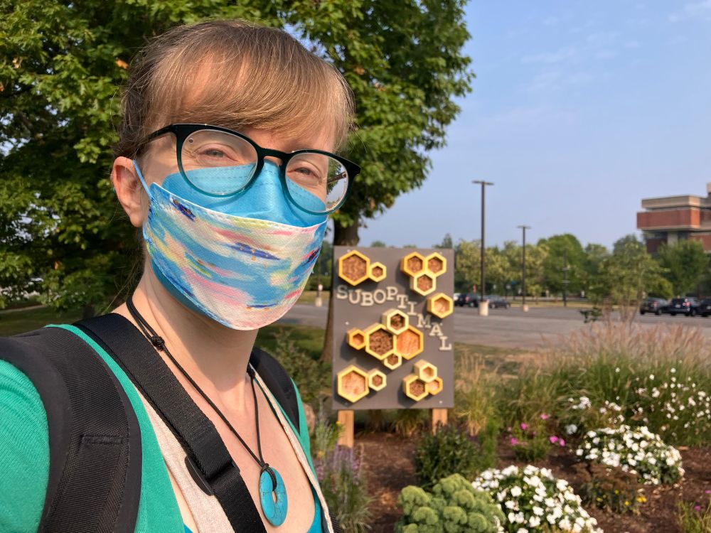 A selfie (dark blonde, white, glasses, wearing a mask because bad air) in front of an outdoor art installation with honeycomb shapes and the word “suboptimal”. 