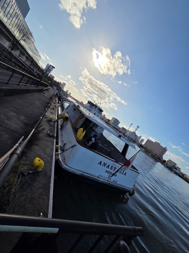 岸壁に係留された白い水上バス船「ANASTASIA NIIGATA」が、静かな水面に浮かんでいる。午後の日差しが雲越しに差し込み、周囲のビルや青空を明るく照らしている。
The white boat “ANASTASIA NIIGATA” is moored along the quay, floating on calm water. Afternoon sunlight shines through the clouds, illuminating the surrounding buildings and the blue sky.