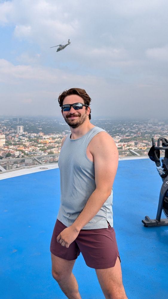 Photo of guy in a grey tank and maroon shirts on top of a helipad in Mexico City, behind him is a spin machine and a helicopter in the sky