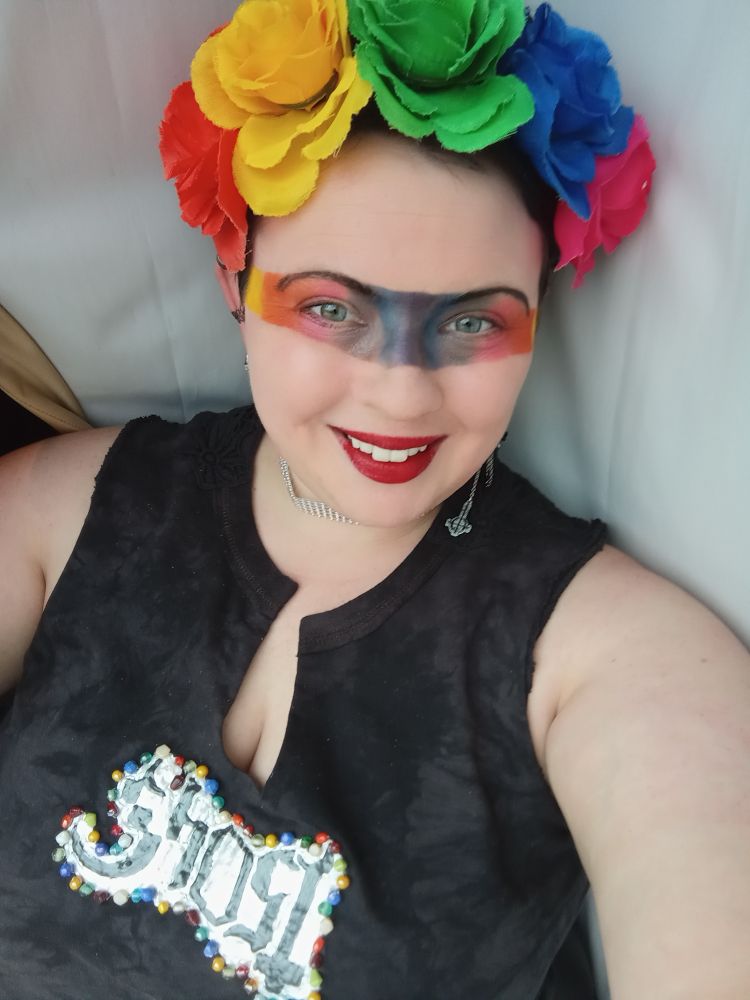 A woman looks at the camera smiling. She has a black crop top that has the band ghost logo on it with glass rainbow beads bordering it. She also wears very dramatic rainbow eyeshadow, and a rainbow flower crown.