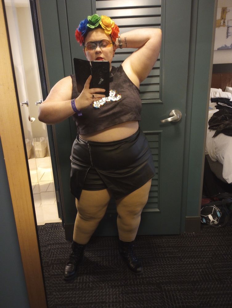 Image description, a woman looks at a hotel mirror showing off their outfit. She wears a leather skirt with built-in shorts, and a acid died black crop top that has the band ghost logo embroidered with rainbow glass beads. She also wears a flower crown and has very dramatic rainbow eyeshadow