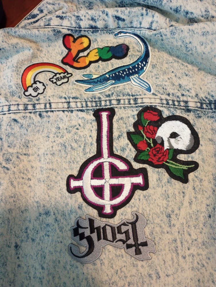 Image description, a close up of the patches on a battle jacket. One of the patches in the shoulder area include a patch that reads love, and rainbow colors. Another patch is a rainbow with two clouds at the ends, with the words oh yeah in them. The other patch is a blue and white plesiosaurus. On the upper back portion is a grucifix that is white with a purple and black outline. A silver patch in Black lettering reads ghost below it. And then the other patch is a Phantom Opera patch with the white half mask and three roses next to it.