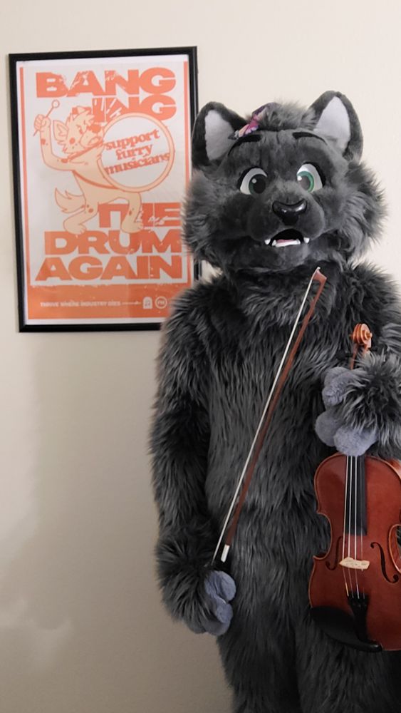 Gray cat fursuit holding a violin standing next to a poster that reads, "BANGING THE DRUM AGAIN" with a hyena banging a drum that reads, "support furry musicians"