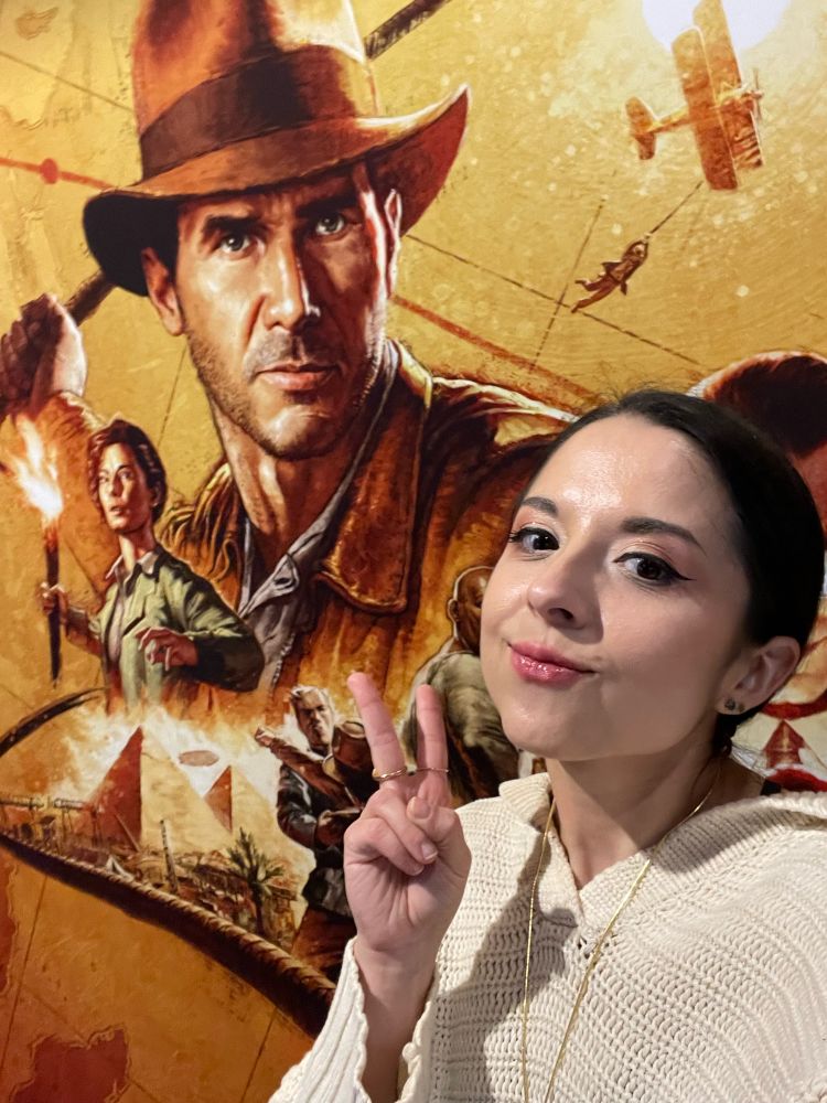 A young woman poses with peace sign fingers in front of a poster for Indiana Jones and the Great Circle.