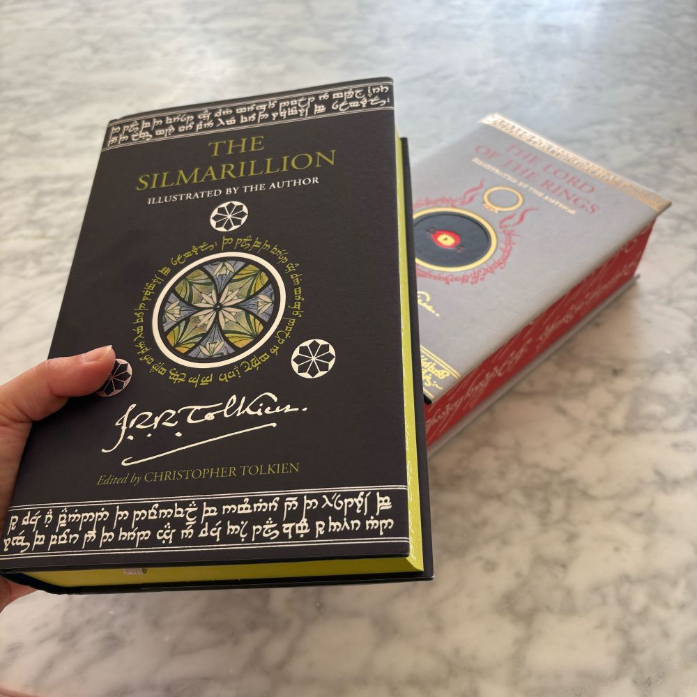 A hand holds a collector’s illustrated edition of The Simarillion by J.R.R. Tolkien. Below it sits on a table a collector’s illustrated edition of The Lord of the Rings by J.R.R. Tolkien.