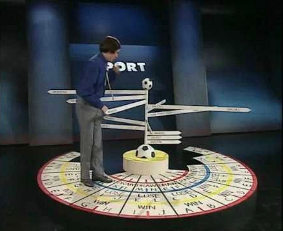 The character Alan Partridge stands on a circular game board in front of a blue backdrop with the word "SPORT" visible, next to a multi-directional signpost with the text, "DRAW", "WIN", and "LOSE" visible. The signpost has two soccer balls attached and is centered on a yellow podium. The circular board reads "DRAW", "WIN", "LOSE", and has sections marked "A" to "M".
