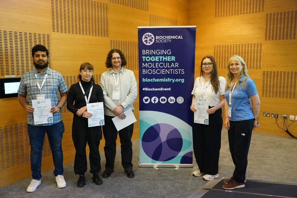 Cameron standing with other prize awardees by a Biochemical Society poster which says bringing together molecular scientists.