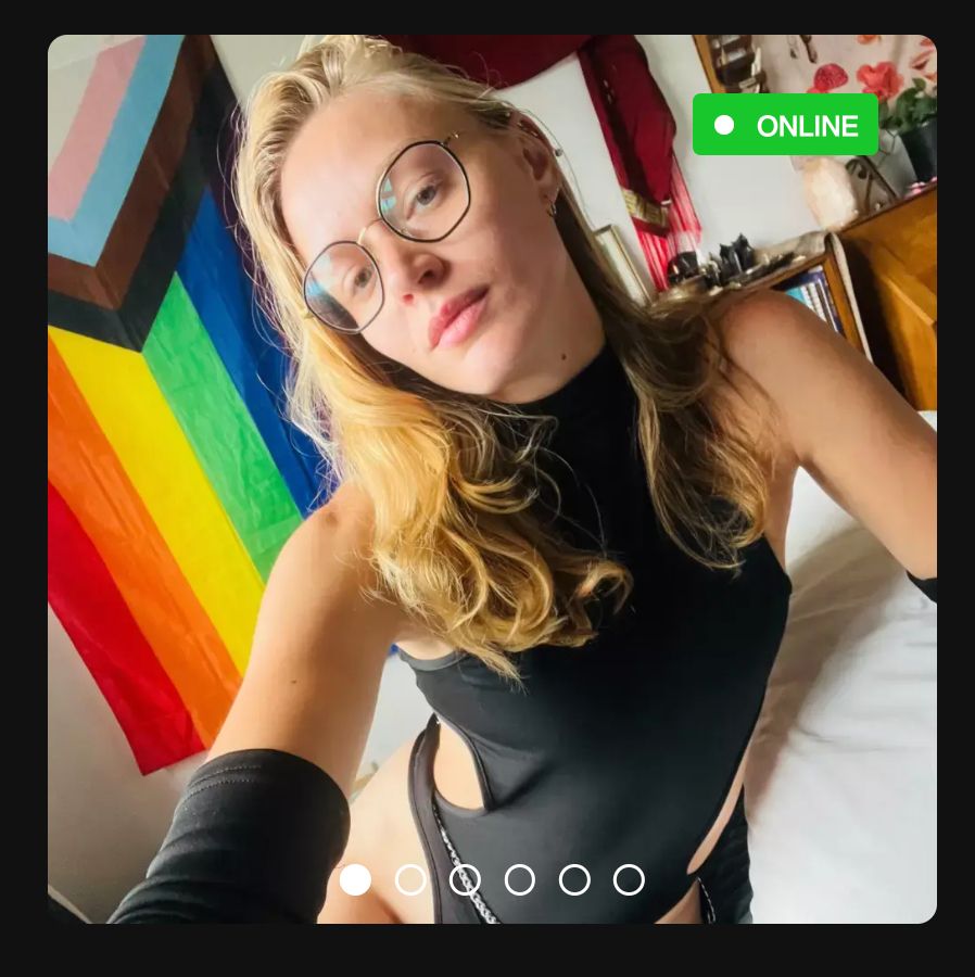 A preview of Jasper's SextPanther profile picture. They're wearing a black bodysuit with round waist cutouts and their blonde hair is cascading down their pecs. A rainbow flag is on the wall behind them, and there's a green rectangle in the top right that says ONLINE. There are round buttons at the bottom, indicating there are more photos to view. Aren't you curious?