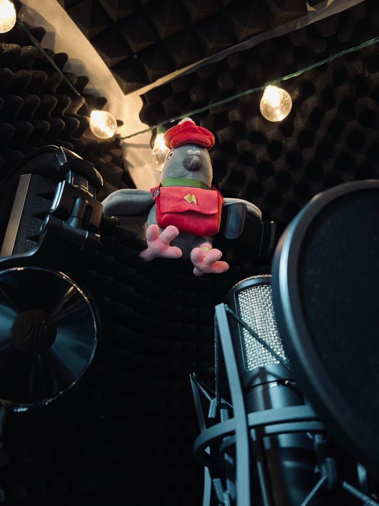 Voice acting booth setup with a mic, pop filter and headphones resting on an isolation shield - a pigeon plushie with a red cap and bag around his neck is perched on top  