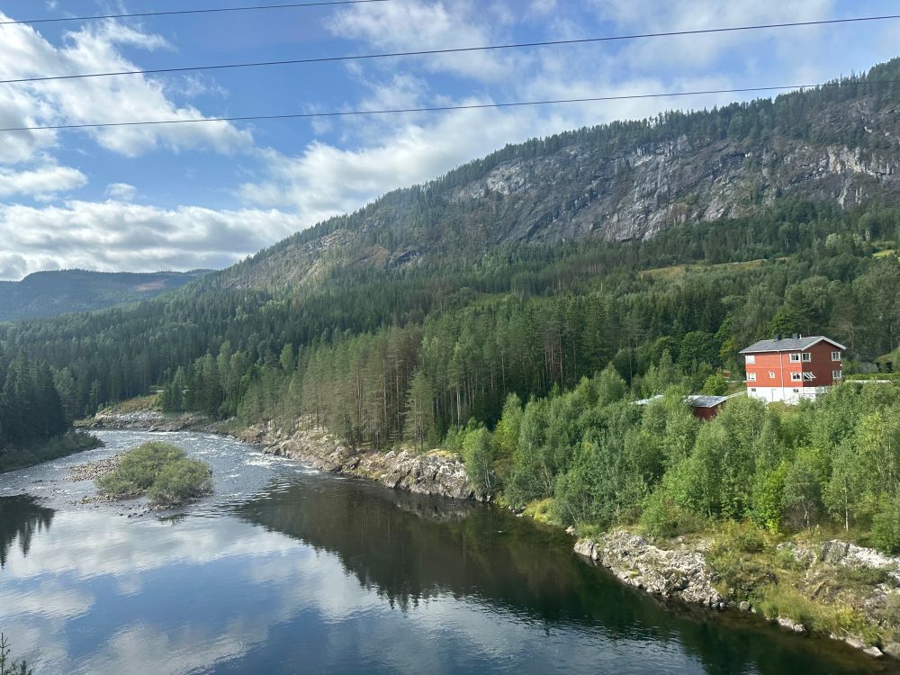 A pretty red building over a rover reflecting the trees and sky overloading by rugged hills.